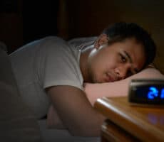 A man is lying in bed with eyes wide open, unable to sleep; in the foreground, a digital clock reads 2:00.