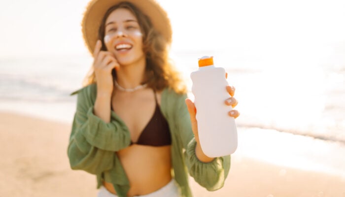 Person wearing a sun hat holding an unbranded sunscreen bottle while standing on a sunny beach.