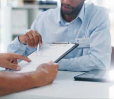 Two people sitting opposite each other across a desk reviewing paperwork on a clipboard.