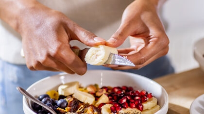 Person cutting slices of banana into bowl of fruit