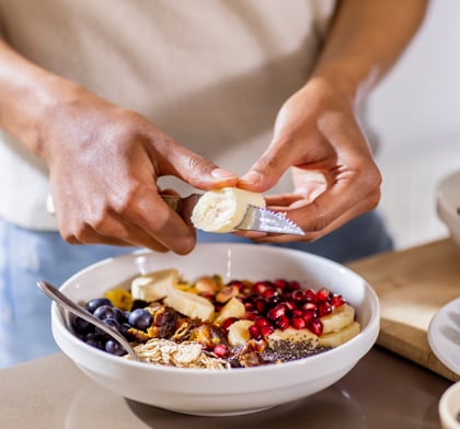 Person cutting slices of banana into bowl of fruit