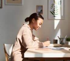 A woman sits at her desk, writing in a journal, in a sunny home office space.