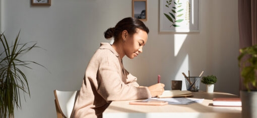 A woman sits at her desk, writing in a journal, in a sunny home office space.