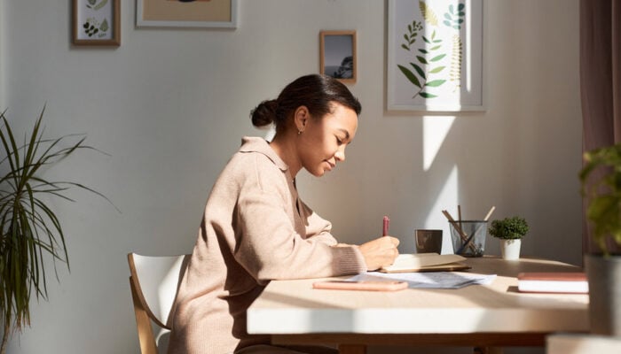 A woman sits at her desk, writing in a journal, in a sunny home office space.