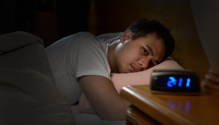 A man is lying in bed with eyes wide open, unable to sleep; in the foreground, a digital clock reads 2:00.