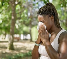 Person outdoors in a park sneezing into a tissue, illustrating allergy symptoms like hay fever