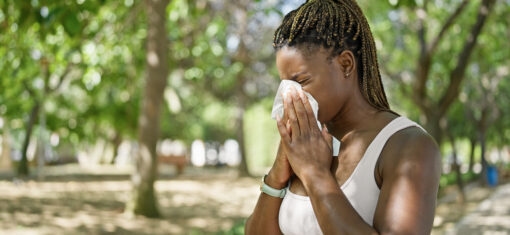 Person outdoors in a park sneezing into a tissue, illustrating allergy symptoms like hay fever
