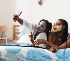 Two women relaxing on a bed wearing face masks and taking a selfie, representing skincare routines and trends