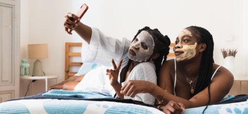 Two women relaxing on a bed wearing face masks and taking a selfie, representing skincare routines and trends