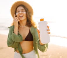 Person wearing a sun hat holding an unbranded sunscreen bottle while standing on a sunny beach.