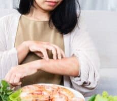 Woman scratching a red rash on her forearm while sitting near a plate of shrimp and fresh greens.