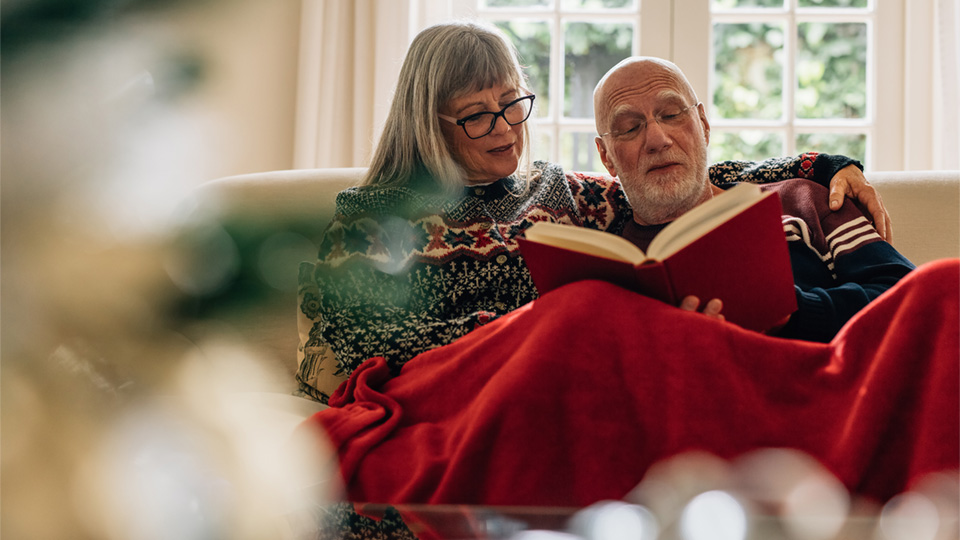 Older man and woman sitting close together on couch under a blanket reading a book