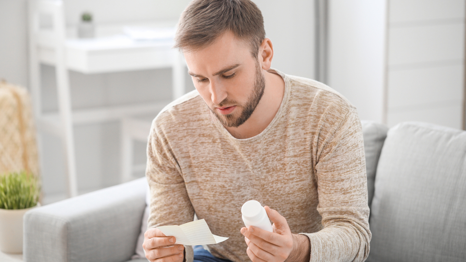 A young man sits in a bright room holding a medication bottle and a prescription paper. He is looking at both items in a contemplative manner.