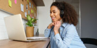 A woman in a denim jacket and headphones sits at her desk watching her laptop.