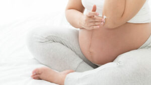 A close up of a pregnant woman wearing gray pants sitting cross-legged on a bed applying white cream to the skin on her elbow.