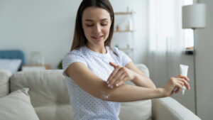 Woman sit on sofa in living room holding small tube with serum caring about skin beauty health care apply ultra-softening cream on elbow area, hydrate nourish it. Beauty treatment and skincare
