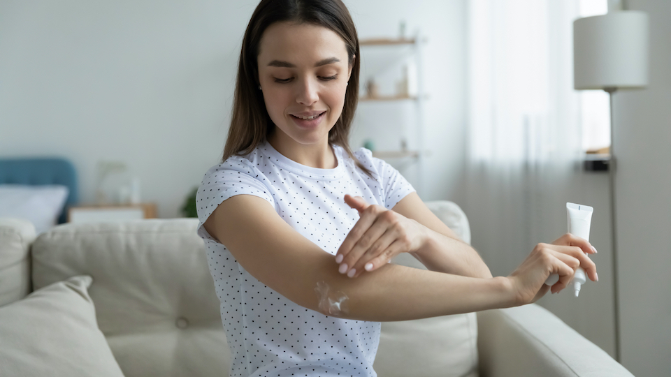 Woman sit on sofa in living room holding small tube with serum caring about skin beauty health care apply ultra-softening cream on elbow area, hydrate nourish it. Beauty treatment and skincare