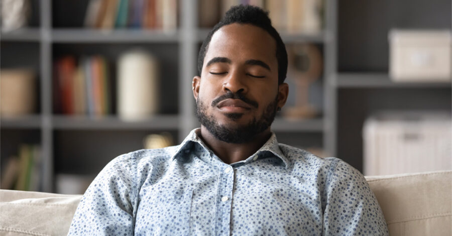 Black man sits on couch in calm therapy setting with eyes closed, soft smile.