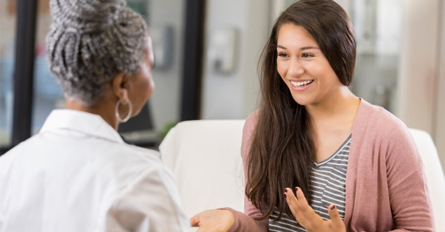 Ethnically ambiguous woman in her 30s wearing pink cardigan talking with black female dermatologist