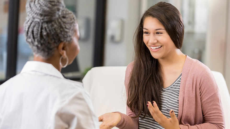 Ethnically ambiguous woman in her 30s wearing pink cardigan talking with black female dermatologist