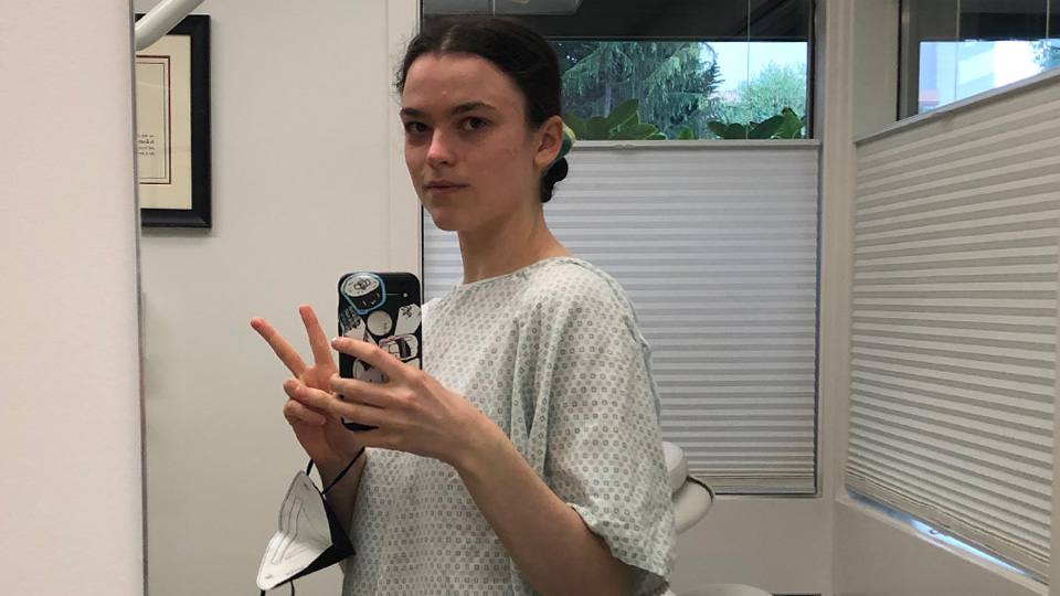 A photo of Karin Stoddard taking a photo of herself in a mirror wearing a medical gown, looking at the camera, and holding up a peace sign with her fingers.