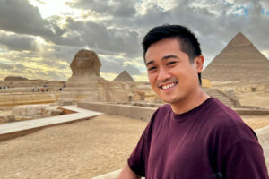 Bryan, a young man with eczema, poses smiling with the Sphinx and an Egyptian pyramid behind him.